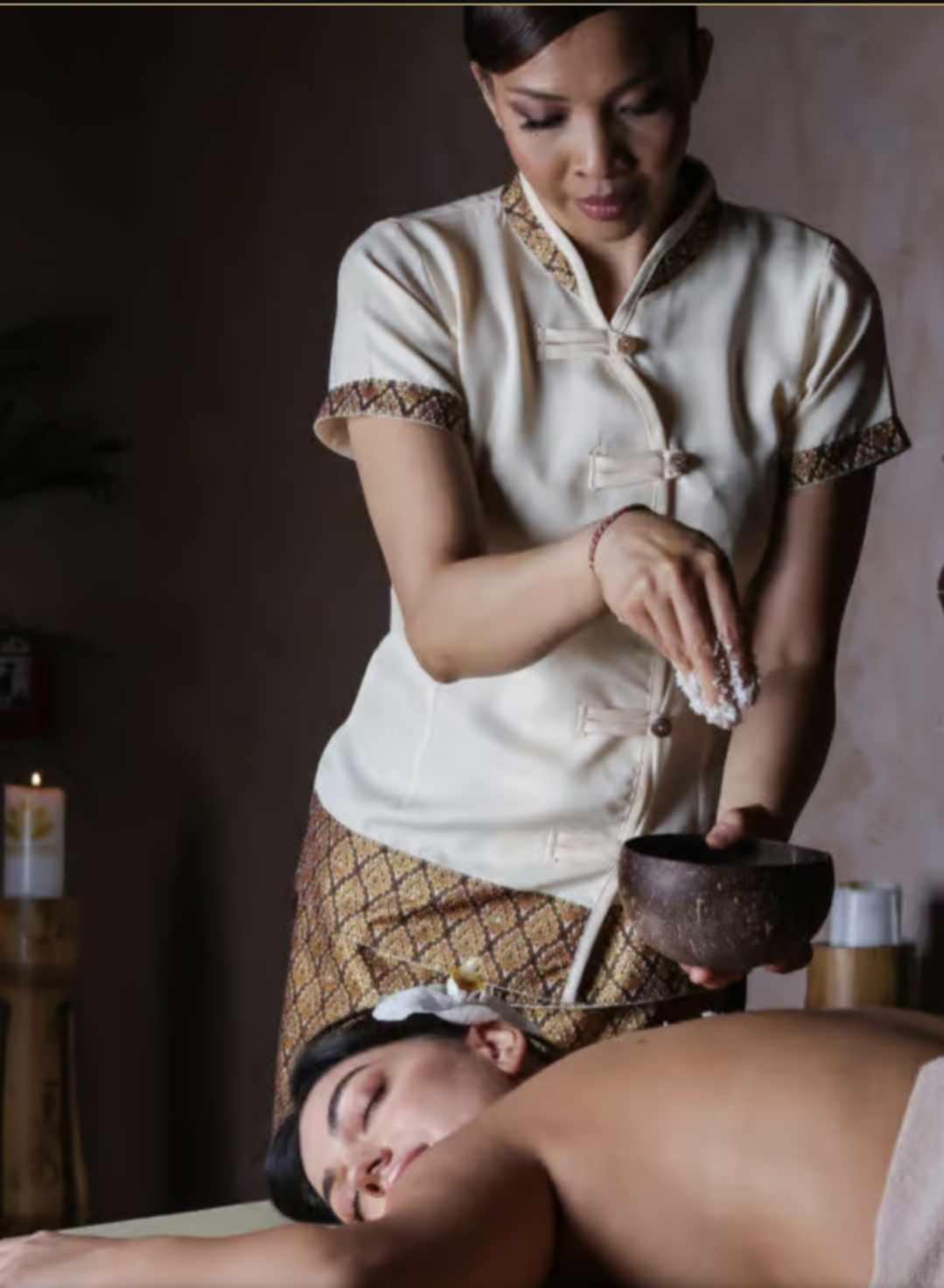 A Nuad Thai therapist in traditional uniform performing a body scrub ritual with a coconut bowl for a relaxed guest