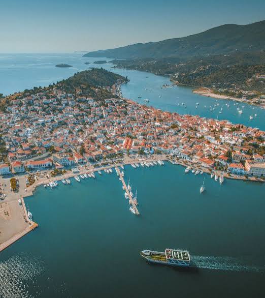Aerial view of Poros island, its red-roofed town between two calm bays and the ferry crossing — a classic Saronic gulf day trip from Athens