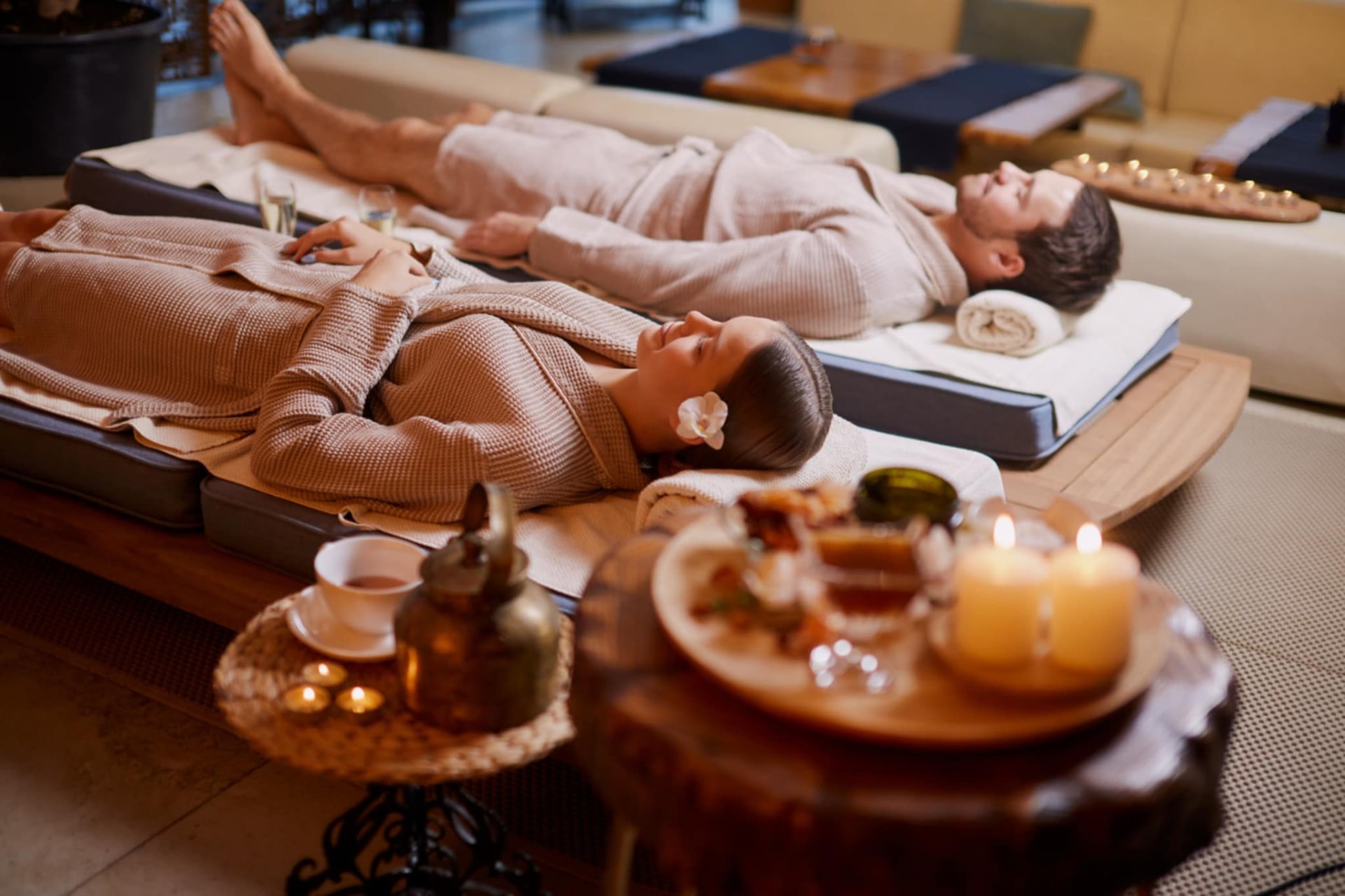 Guests in robes resting on the raised mattresses of a traditional Thai massage room, with herbal tea and candles, at Nuad Thai Massage Athens