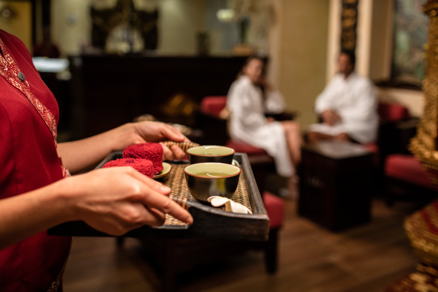 Thai therapist in red uniform carrying a tray of herbal tea, rolled towels and fresh petals for guests