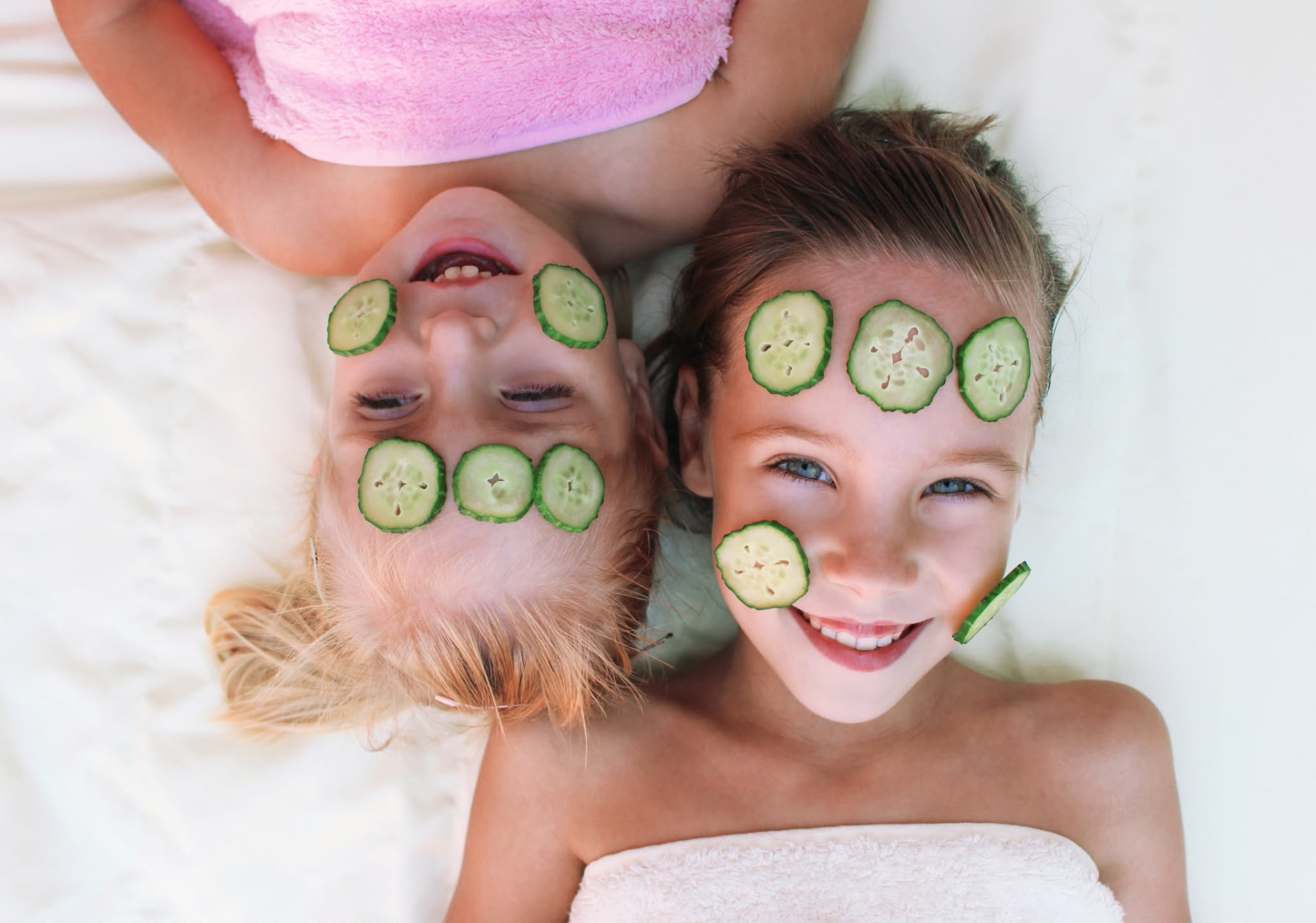 Two smiling children wrapped in towels with cooling cucumber slices on their faces, enjoying a kids spa day at Nuad Thai Massage Athens
